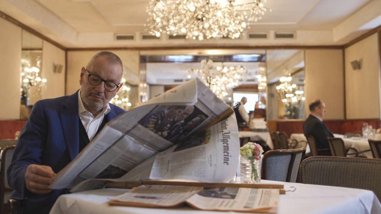 Das Bild zeigt eine elegante Café-Umgebung. Ein Mann sitzt an einem Tisch und liest eine aufgeschlagene Zeitung. Er trägt eine Brille und ein helles Hemd unter einer dunkelblauen Jacke. Auf dem Tisch liegen weitere Zeitungen, und ein Glas Wasser steht bereit. Im Hintergrund sind andere Gäste sichtbar, darunter ein Mann, der alleine an einem Tisch sitzt. Die Wände des Cafés sind hell mit Spiegeln dekoriert, und es gibt mehrere funkelnde Kronleuchter, die für eine gehobene Atmosphäre sorgen. Der Raum wirkt gut beleuchtet und einladend.
