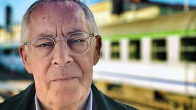 Porträtfoto eines älteren Mannes mit Brille in einem Bahnhof. Im Hintergrund sind Bahngleise und ein Zug zu sehen.
