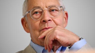 Porträtfoto eines älteren Mannes mit Brille, der nachdenklich eine Hand an den Mund legt.