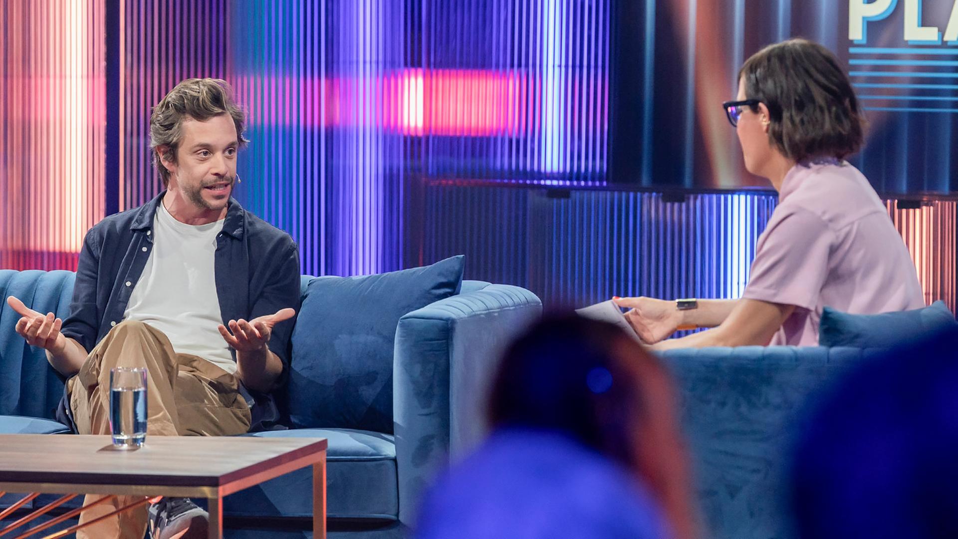 Eva Karl Faltermeier und ihr Gast, Tobias Krell (Checker Tobi), unterhalten sich auf blauen Sofas im Studio.