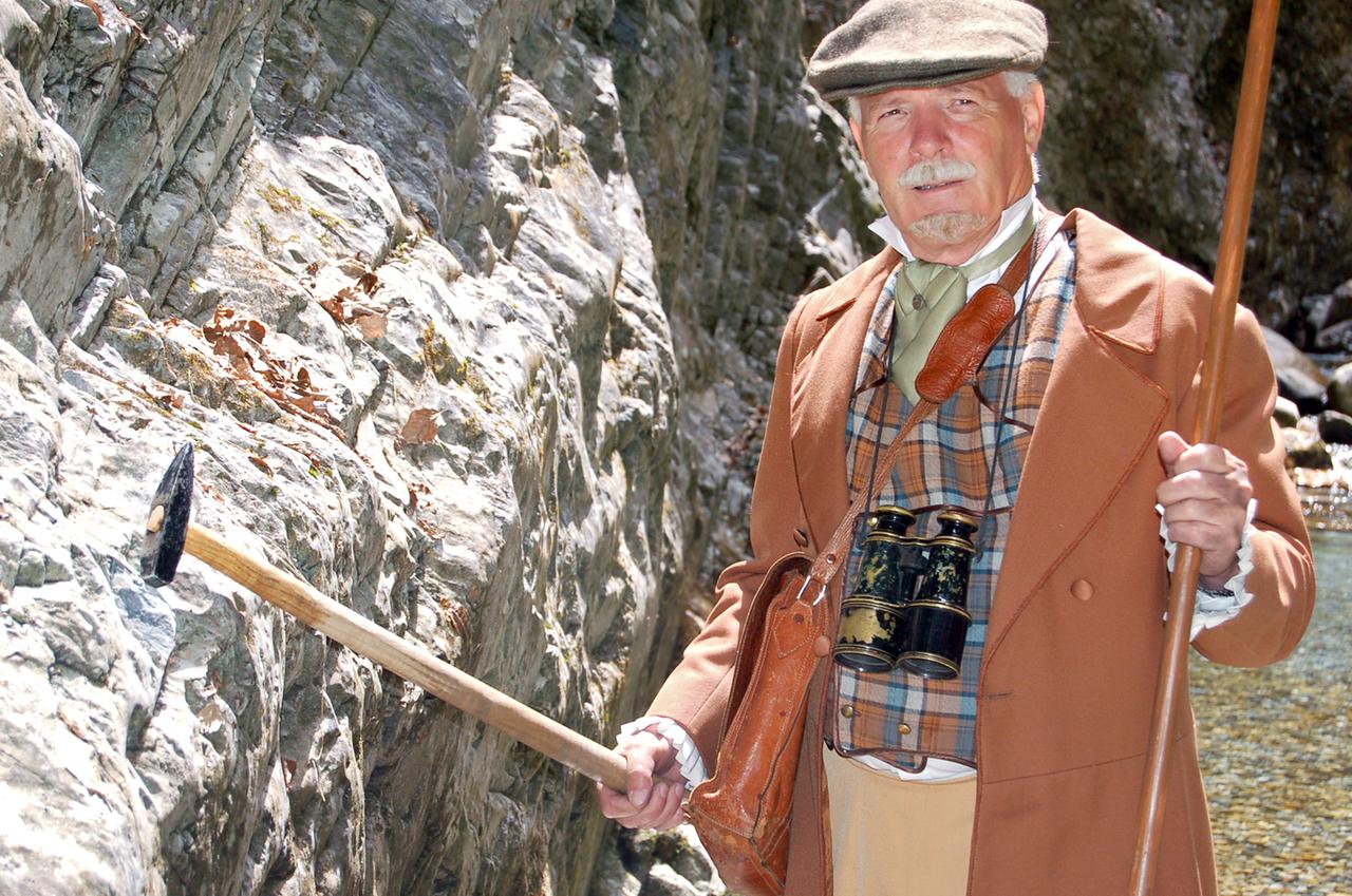 Ein historischer Geologe steht vor einer Felswand mit einem Hammer.