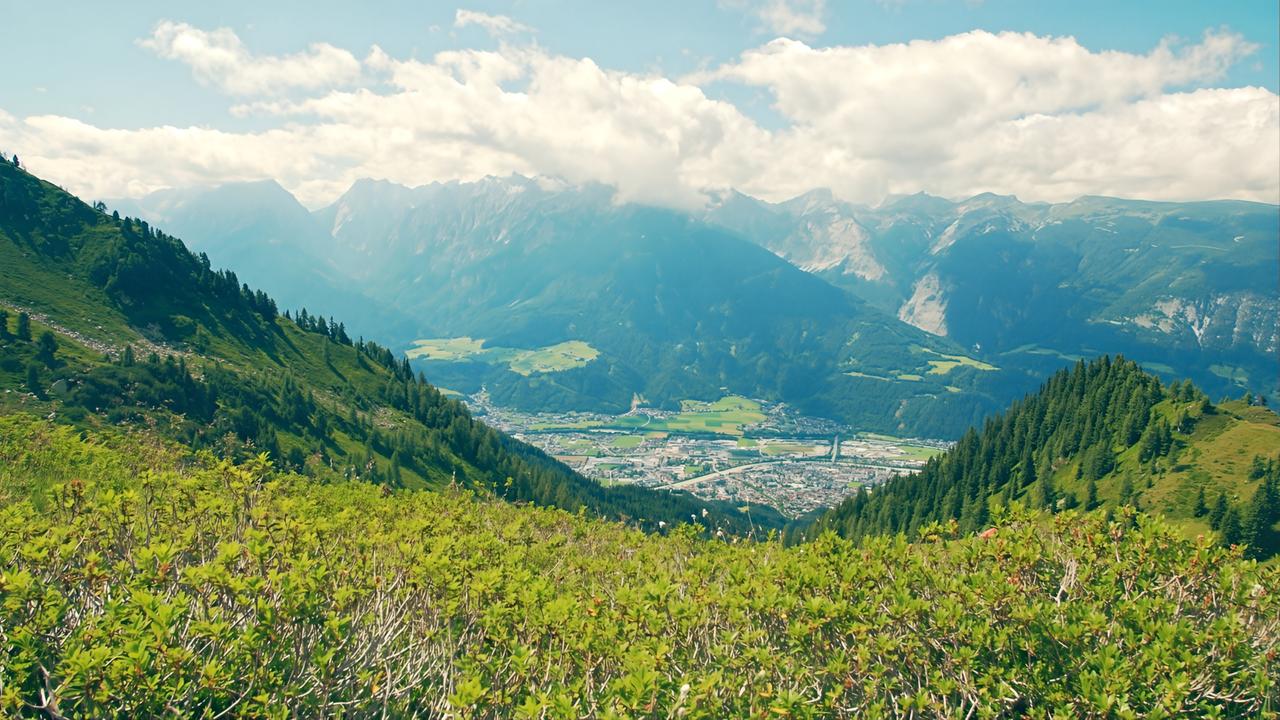 Das Bild zeigt eine beeindruckende Aussicht vom Gipfel auf das Tal, das von sanften Hügeln und bewaldeten Bergen umgeben ist. Im Vordergrund sind grüne Sträucher und Pflanzen zu sehen, die den Vordergrund bilden. Die Landschaft im Hintergrund wird von den majestätischen Gipfeln der Berge dominiert, die teils bewaldet und teils schroff sind. Einige Berge sind von Wolken umgeben, was dem Bild eine mystische Atmosphäre verleiht.  Unten im Tal erkennt man die Stadt Schwaz, die von landwirtschaftlichen Flächen und einigen Gebäuden umgeben ist. Die Stadt ist gut in die grüne Umgebung integriert, mit einer klaren Sicht auf die Straßen und Strukturen. Der Himmel ist helle und hat einige Wolken. Insgesamt vermittelt die Darstellung ein Gefühl von Weite und der Schönheit der Tiroler Natur, sowie der harmonischen Verbindung zwischen Stadt und Landschaft.