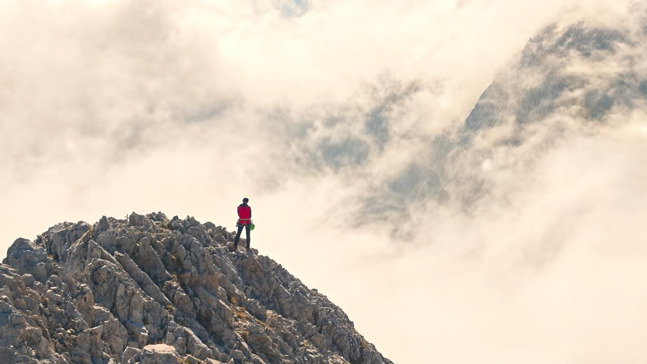 Das Bild zeigt eine hochgelegene, zerklüftete Berglandschaft im Karwendelgebirge. In der Mitte des Bildes steht eine Person in einem roten Shirt, die auf einem Felsen steht und in die Ferne blickt. Der Hintergrund ist von dichten, weißen Wolken und Nebel geprägt, die einen dramatischen Kontrast zu den harten Felsstrukturen bilden. Die Person hat einen Rucksack auf und scheint die Aussicht zu genießen. Um sie herum sind weitere, schroffe Felspartien sehr sichtbar. Die Szenerie vermittelt ein Gefühl von Höhe und Weite, typisch für alpine Landschaften.