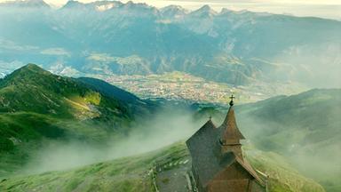 Das Bild zeigt die Kellerjochkapelle, die sich auf einem Bergvorsprung im Karwendelgebirge befindet. Die Kapelle hat einen traditionellen, schindelgedeckten Dachstuhl und ist mit einem kleinen Kreuz auf der Spitze versehen. Vor der Kapelle erstreckt sich eine grüne, hügelige Landschaft, die sanft abfällt und in die darunter liegende Stadt Schwaz übergeht. Im Hintergrund sieht man die majestätischen Gipfel der umliegenden Berge, die teilweise in Nebel gehüllt sind. Die Landschaft ist von einer dichten Vegetation geprägt, die zwischen Wald- und Wiesengebieten wechselt. Am Fuß des Berges sind die Strukturen der Stadt Schwaz und landwirtschaftliche Flächen zu erkennen, die sich in der Talsohle ausbreiten. Die gesamte Szene vermittelt ein Gefühl von Ruhe und Erhabenheit, während die Wolken und der Nebel eine mystische Atmosphäre schaffen.