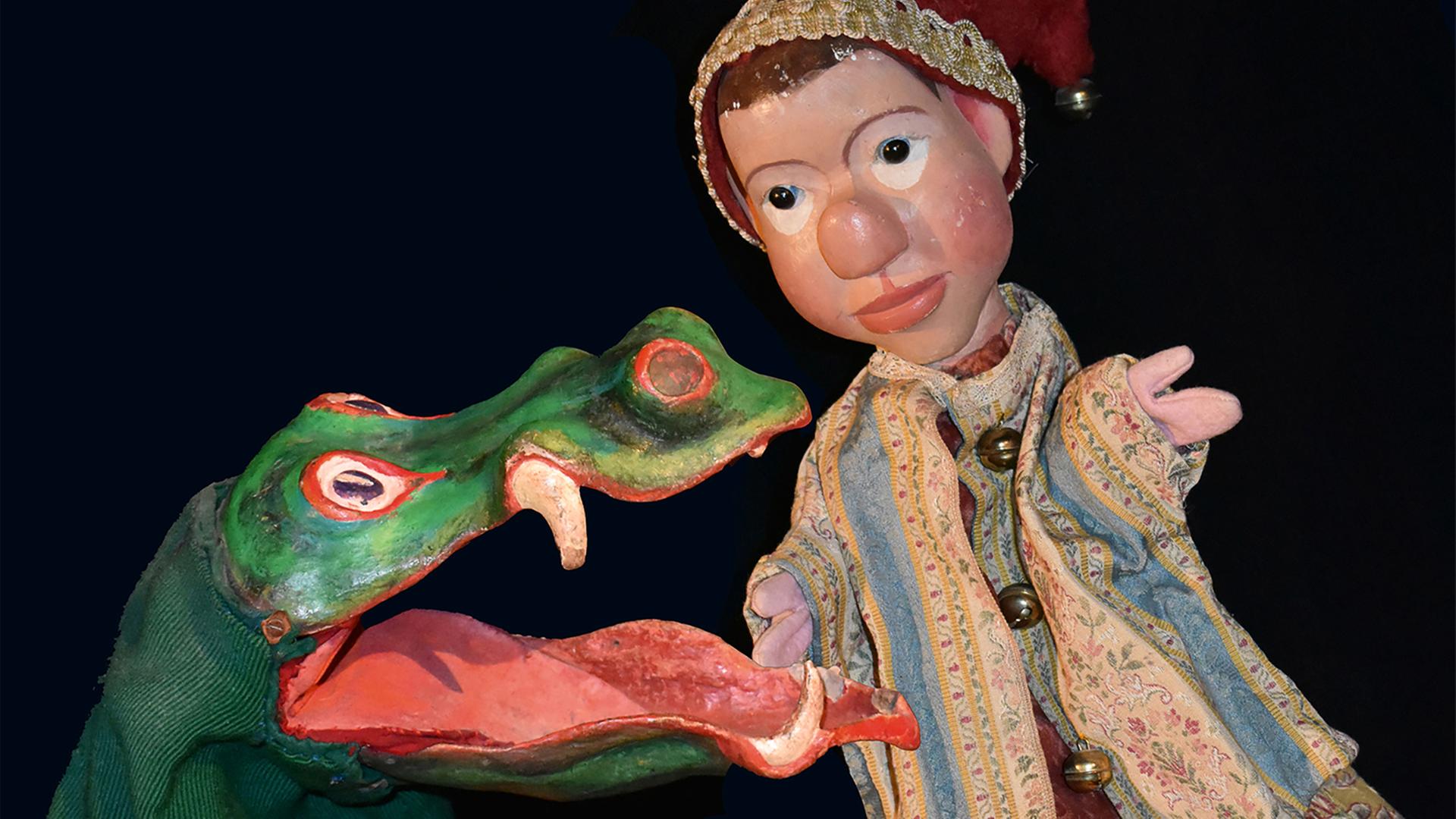 Links ist eine Krokodilpuppe zu sehen und daneben eine Kasperfigur, also ein Junge, der eine rote Zipfelmütze mit goldenem Glöckchen an der Spitze trägt.