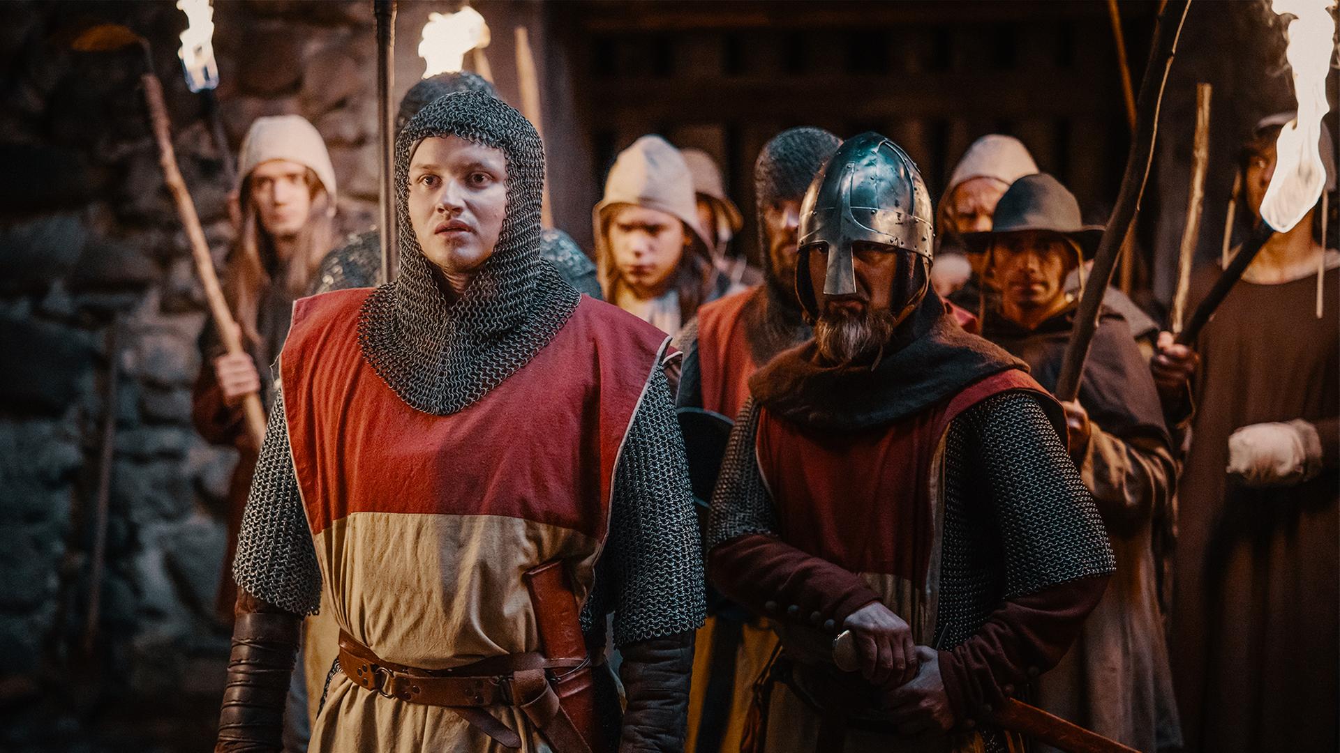 Eberhard von Münzenberg (Paul Lux) steht in seiner Ritterkleidung vor den Mitgliedern der Burgwache in einem Gang. Fackeln brennen.