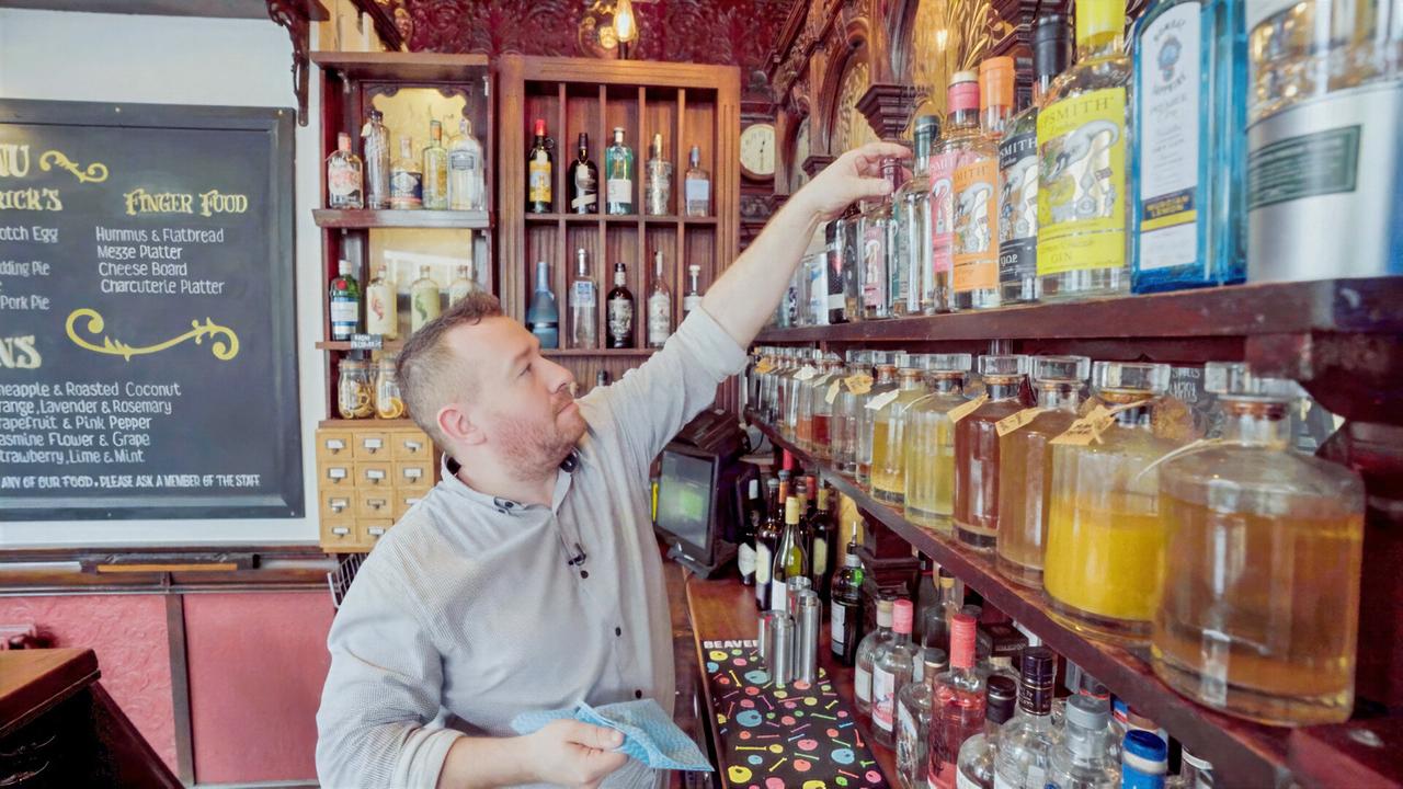 Das Bild zeigt einen Pub-Manager, der in einer traditionellen Bar arbeitet. Er steht vor einer Holzregalwand, auf der eine Vielzahl von Spirituosen und Flaschen angeordnet sind. Der Mann hat einen kurzen Bart und trägt ein helles, langärmliges Hemd. Er greift mit der rechten Hand nach einer Flasche, während er mit der linken Hand ein Reinigungstuch hält. Hinter ihm ist eine große Tafel angebracht, auf der Speisen aufgelistet sind, und es sind auch einige kommentierende Texte in geschwungener Schrift sichtbar. Der Raum ist gemütlich und warm, mit dunklem Holz und dekorativen Elementen. Die Atmosphäre wird durch das warme Licht, das von hängenden Lampen ausgeht, unterstrichen.