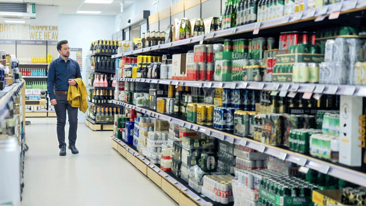 Das Bild zeigt einen Innenraum eines Geschäfts, das auf den Verkauf von alkoholischen Getränken spezialisiert ist. In der Mitte des Raumes sieht man einen Mann, der einen gelben Pullover über dem Arm trägt, während er durch den Gang geht. Er trägt ein dunkelblaues Hemd und graue Hosen. Um ihn herum sind Regale, die mit verschiedenen alkoholischen Getränken gefüllt sind, darunter Bierdosen, Weinflaschen und andere alkoholhaltige Produkte.   Die Regale auf der linken Seite sind unterschiedlich gefüllt und zeigen eine Vielzahl von Getränken in verschiedenen Verpackungen und Farben. Die Beleuchtung im Raum ist hell und sorgt für eine klare Sicht auf die Waren. An der Wand im Hintergrund sind einige Wörter in großen Buchstaben zu sehen, die möglicherweise eine Beschreibung oder eine Werbung darstellen, allerdings sind sie aufgrund der Bildperspektive nicht ganz klar lesbar.   Die gesamte Atmosphäre wirkt ordentlich und organisiert, mit einer klaren Präsentation der Artikel.