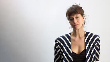 Foto einer jungen Frau mit hochgesteckten Haaren, die eine schräg schwarz-weiß gestreifte Jacke über einem schwarzen Shirt und einer schwarzen Hose trägt.