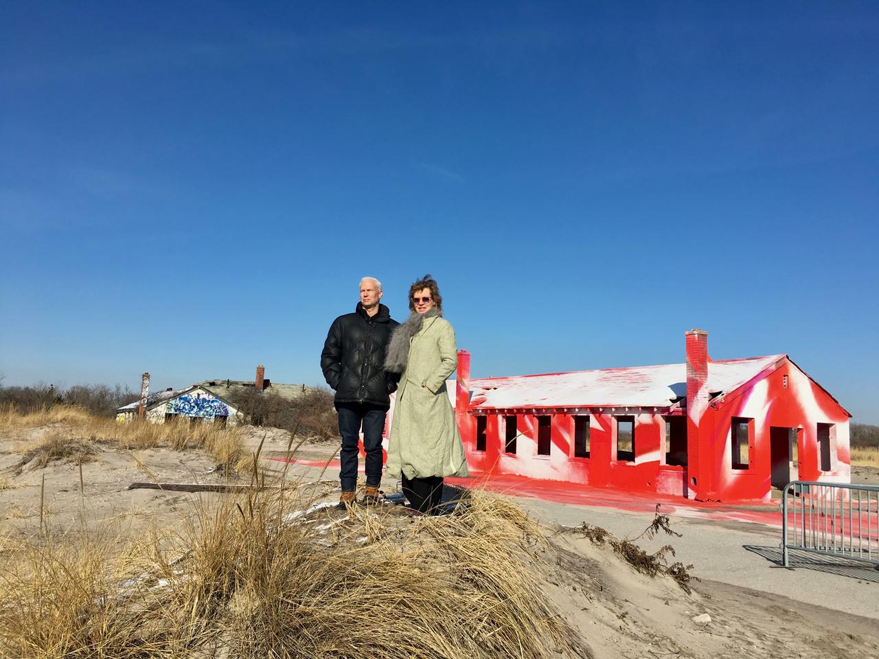 Eine Frau mit hellem Mantel, Sonnenbrille und Schal und ein dunkelgekleideter Mann mit kurzen weißen Haaren stehen auf Sand in der Ruine eines Hauses, das von innen rot und weiß bemalt wurde. 