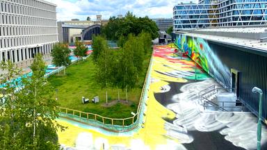 Katharina Grosse Aussenarbeit Hamburger Bahnhof: Blick auf verschiedene Industriehallen und Bürogebäude. In der Mitte eine Grünfläche mit kleinen Birken, drum herum ist der Boden großflächig bunt bemalt, ebenso wie die Außenwand einer Halle am rechten Bildrand. 