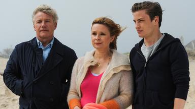 Drei Personen stehen am Strand und blicken in die Ferne. Eine Frau mit blondem Haar trägt eine beige Strickjacke über einem pinken Oberteil, neben ihr ein junger Mann in dunkler Kapuzenjacke und ein älterer Mann in dunklem Mantel.