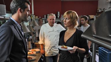 In einer Restaurantküche stehen sich ein Mann im Anzug und eine Frau in schwarzer Kleidung gegenüber. Sie hält einen Teller mit einem Gericht, während im Hintergrund ein Koch und weitere Personen zu sehen sind.