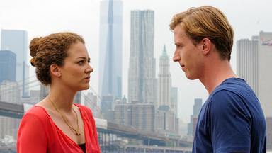 Eine Frau und ein Mann stehen sich an einem Flussufer gegenüber, mit der Skyline von Manhattan im Hintergrund. Die Frau trägt eine rote Bluse, der Mann ein blaues T-Shirt.