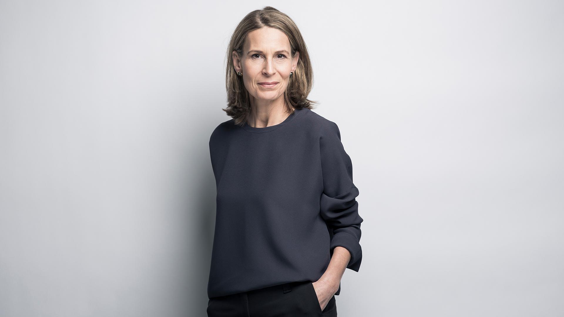 Eine Frau mit mittelblonden Haaren steht vor einer hellen Wand.