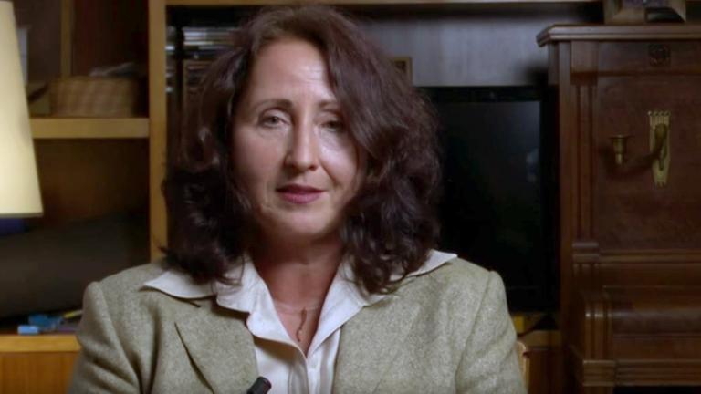 Ein Frau mit braunen Haaren sitzt vor einem dunklen Schrank und schaut während einer Gesprächsituation in die Kamera.