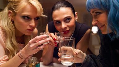 Das Bild zeigt drei Frauen, die an einem Tisch sitzen und kleine Gläser mit einem klaren Getränk halten. Die Frauen haben verschiedene Haarfarben und Frisuren.   Die erste Frau links hat lange, blonde Haare und wirkt konzentriert; sie trägt ein zartes, helles Oberteil und Schmuck. Die zweite Frau in der Mitte hat dunkle, glatte Haare und trägt ein schwarzes, elegantes Kleid; sie hat einen ernsten Gesichtsausdruck, während sie einem anderen ins Auge schaut. Die dritte Frau rechts hat kürzere, türkisfarbene Haare und trägt ein karierter Hemd; sie lächelt und schaut ebenfalls auf das Glas.  Auf dem Tisch sind auch einige Snacks zu sehen, wie Gurke und Wurst, sowie ein Glas mit einem klaren Getränk. Der Hintergrund des Bildes ist unscharf, aber es scheinen Möbel oder weitere Gegenstände zu sein. Die Szene vermittelt eine Atmosphäre der Anspannung und des Austauschs zwischen den Frauen.