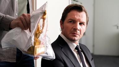 Ein Mann sitzt auf einem Stuhl und betrachtet eine in eine Plastiktüte verpackte Statue, die von einer Hand gehalten wird.