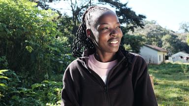 Eine junge Kenianerin sitzt in der Natur. Im Hintergrund sind Büsche, eine Wiese und Hütten zu sehen.