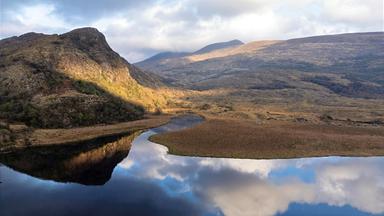 Blick über eine Moorlandschaft auf der Halbinsel Kerry in Irland mit einem dunklen, geschwungenen See im Vordergrund, felsigen Bergen und dicht bewölktem Himmel.