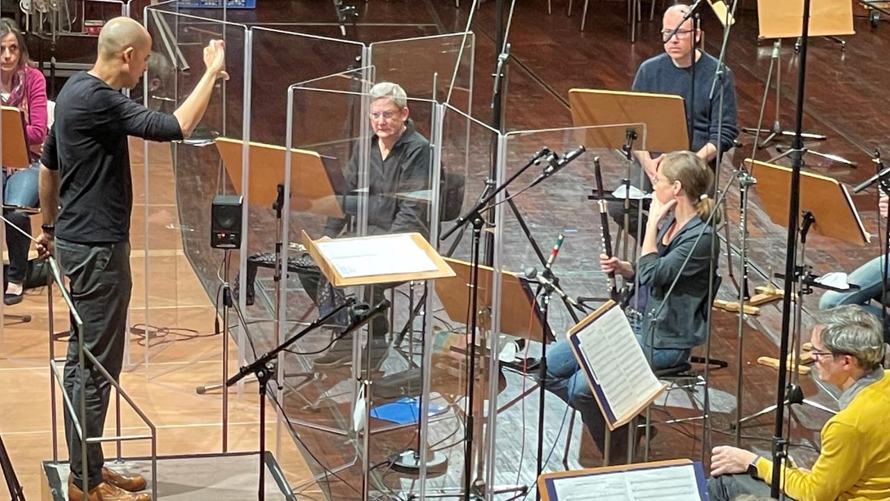 Der Schwarze Dirigent Kevin John Edusei dirigiert weiße Orchestermusiker hinter Plexiglasabtrennungen.