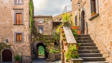 Rustikale Gasse in einer alten europäischen Stadt mit steinernen Gebäuden, begrünten Wänden, einer Treppe mit Blumentöpfen und einem Blick durch einen Torbogen auf eine grüne Landschaft.