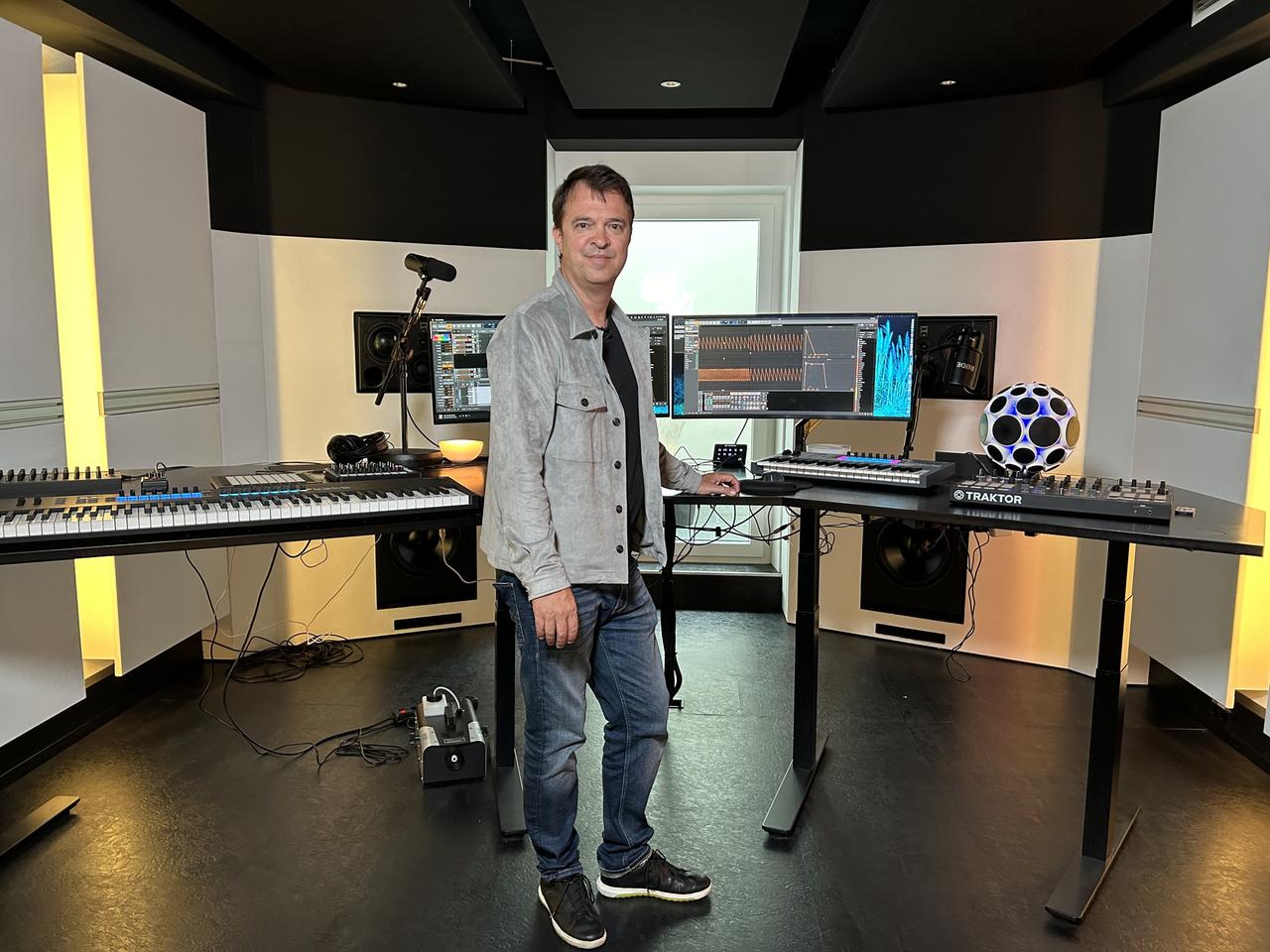Mann mit kurzen Haaren in Jeans und Jeansjacke steht in Tonstudio, links ein Keyboard, in der Mitte und recht Computer und Monitore.