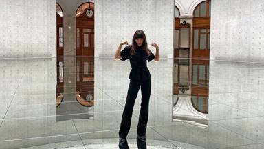  In einem runden Raum mit hohen Betonwänden und einem spiegelnden Boden steht eine Frau mit langen, dunklen Haaren und hebt beide Arme an, als würde sie ihre Muskeln präsentieren. Sie trägt ein komplett schwarzes Outfit, bestehend aus einer taillierten Bluse mit kurzen Ärmeln und einer weiten Hose. Ihr Blick ist selbstbewusst und direkt in die Kamera gerichtet. Der Boden reflektiert die Umgebung, wodurch es scheint, als würde der Raum nach unten hin weitergehen. Durch zwei große, rechteckige Öffnungen in den Betonwänden sind elegante, historische Elemente wie Holzvertäfelungen, Rundbögen und eine große Wanduhr sichtbar.