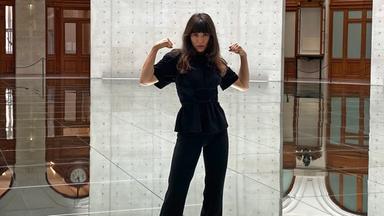  In einem runden Raum mit hohen Betonwänden und einem spiegelnden Boden steht eine Frau mit langen, dunklen Haaren und hebt beide Arme an, als würde sie ihre Muskeln präsentieren. Sie trägt ein komplett schwarzes Outfit, bestehend aus einer taillierten Bluse mit kurzen Ärmeln und einer weiten Hose. Ihr Blick ist selbstbewusst und direkt in die Kamera gerichtet. Der Boden reflektiert die Umgebung, wodurch es scheint, als würde der Raum nach unten hin weitergehen. Durch zwei große, rechteckige Öffnungen in den Betonwänden sind elegante, historische Elemente wie Holzvertäfelungen, Rundbögen und eine große Wanduhr sichtbar.