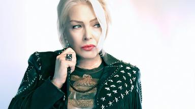 Das Bild zeigt eine weibliche Person mit blonden Haaren, die zu einem modernen, leichten Make-up-Stil gestylt ist. Sie trägt eine schwarze Jacke mit auffälligen, hellen Sternen und Fransen an den Ärmeln. Darüber hat sie ein dunkles Oberteil mit einem goldenen Muster an. Die Person hat einen nachdenklichen Ausdruck und schaut leicht zur Seite, während sie einen Ring an der Hand hält. Der Hintergrund ist hell und unscharf, sodass der Fokus auf der Person liegt. Es handelt sich offenbar um eine Künstlerin, die in einem musikalischen Kontext präsentiert wird, da im Text auf ein Konzert und ihre erfolgreichsten Songs verwiesen wird.