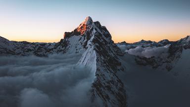 Eine Luftaufnahme des verschneiten, felsigen Berggipfels mit einem steilen Grat. Unterhalb liegen die Wolken.