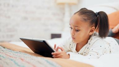 Mädchen liegt allein auf dem Bett und schaut aufs Tablet. 