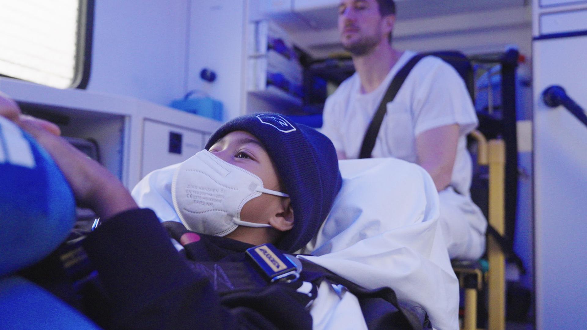 Ein Kind mit FFP2-Maske und Mütze liegt angeschnallt auf einer Trage im Inneren eines Krankenwagens; im Hintergrund sitzt ein Erwachsener mit Schultergurt.