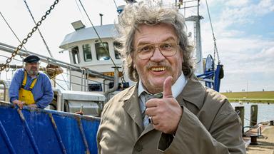 Mann steht auf einem Schiffskutter und reckt den Daumen optimistisch nach oben. Sein Aussehen und sein Gebiss wirken ungepflegt.