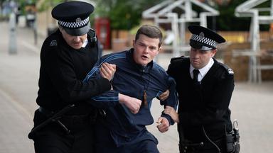 Zwei britische Polizisten in Uniform führen einen jungen Mann Trainingsanzug ab, der sich wehrt.
