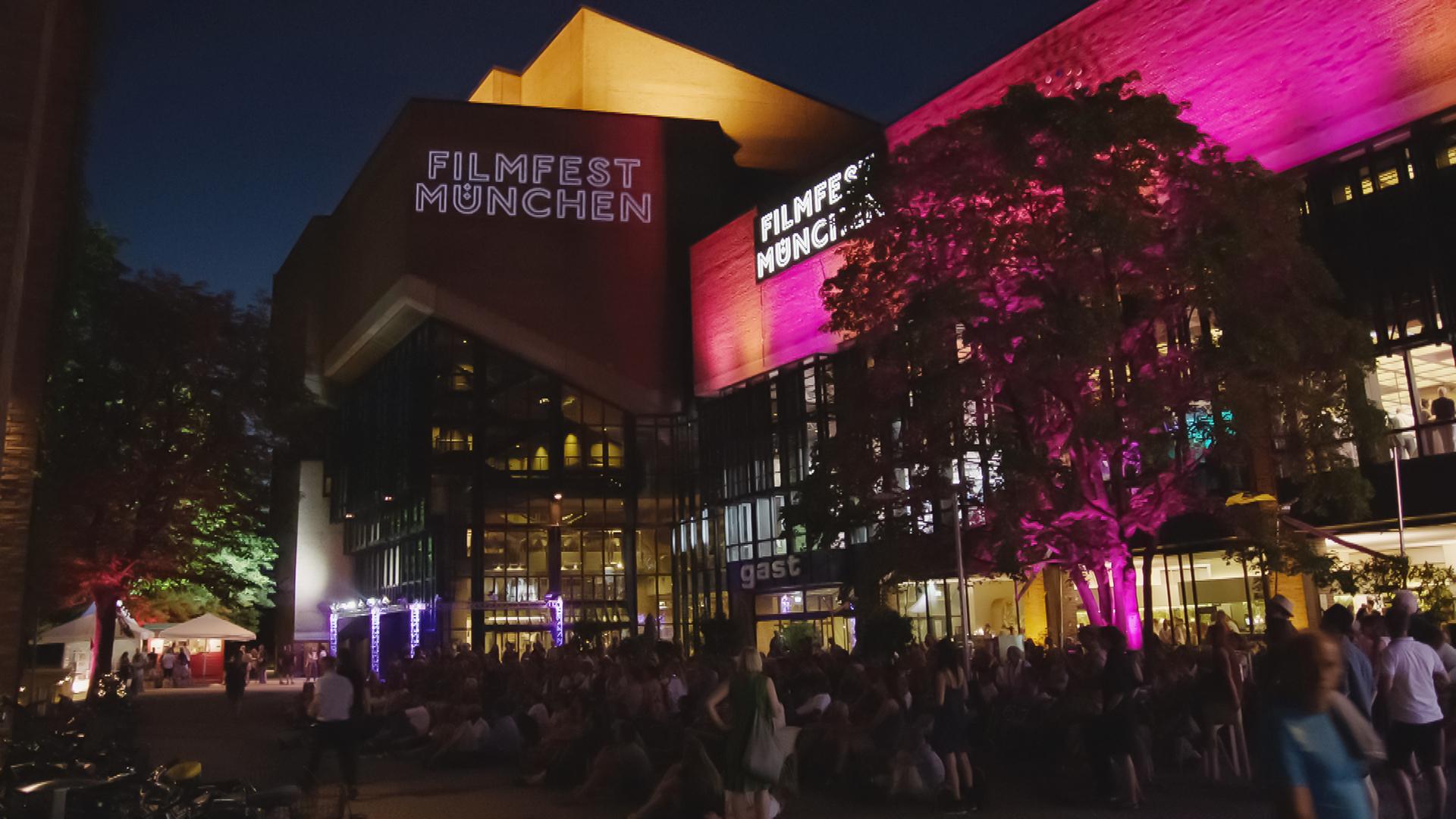 Das gelb und pink beleuchtete Amerikahaus in München mit dem Schriftzug des Filmfests München. Vor dem Haus eine Menschenmenge von Festivalbesuchern.