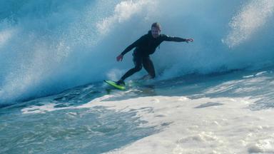 Ein junger Mann im Neoprenanzug steht auf einem Surfbrett und surft auf einer großen Welle, die hinter ihm bricht