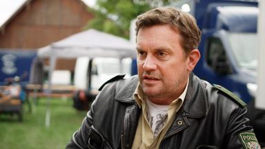 Sebastian Bezzel in Polizei-Lederjacke sitzt vor einem Lieferwagen und einem Zelt.