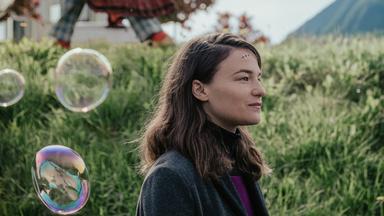 Eine Frau mit einer kleinen, geklammerten Wunde auf der Stirn steht lächelnd vor einem Grashügel.