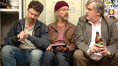 Drei Männer sitzen vor einem Spinnt. Einer hält eine Flasche Bier in der Hand, ein anderer ein Mobiltelefon. Der dritte Mann schaut auf seine Armbanduhr.