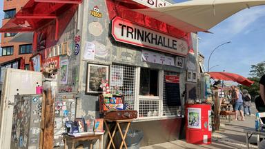 Das Bild zeigt einen Kiosk mit dem Namen „Trinkhalle“, der an einer Straßenecke steht. Über dem Schalter hängt ein Schild mit der Aufschrift „Trinkhalle Seit 1876“. Die Fassade des Kiosks ist abgenutzt und mit zahlreichen Stickern und Graffiti bedeckt. Vor dem Kiosk stehen einige Bücher auf einem kleinen Tisch, daneben verschiedene dekorative Elemente wie Bilder und andere Kunstgegenstände. Rechts im Bild sind einige Menschen unter einem Sonnenschirm zu sehen, die an Tischen stehen. 
