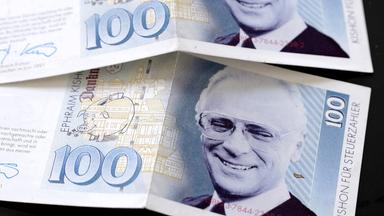 Das Bild zeigt eine 100-Euroschein-Grafik mit dem Porträt von Ephraim Kishon. Der Schein ist gefaltet und liegt auf einer dunklen Oberfläche. Im Hintergrund sind feine grafische Elemente zu erkennen.