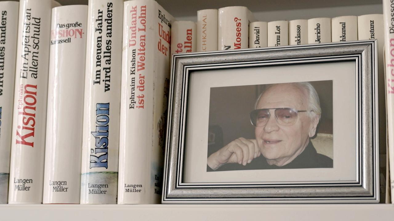 Das Bild zeigt ein Bücherregal mit mehreren Exemplaren von Ephraim Kishons Werken. In einem Rahmen steht ein Foto eines Mannes mit Brille und grauen Haaren, der entspannt lächelt.