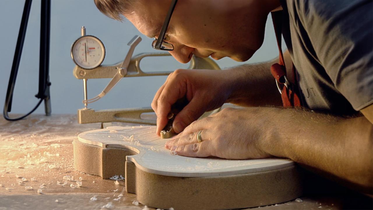 Das Bild zeigt einen Geigenbauer, der konzentriert an einem Geigenkörper arbeitet. Er trägt eine Brille und hat eine Goldring an der linken Hand. Mit einem Werkzeug in der rechten Hand bearbeitet er die Holzoberfläche der Geige, die auf einem Holztisch liegt. Um den Geigenbauer herum sind Holzspäne zu sehen, die sich auf der Tischoberfläche verteilt haben. Im Hintergrund befindet sich ein Messgerät, das offenbar zur Überprüfung der Dimensionen des Instruments dient, sowie eine dunkle Struktur, möglicherweise eine Kamera oder ein weiteres Werkzeug. Das Licht im Raum ist weich und betont die Details der Arbeit und des Holzmaterials.