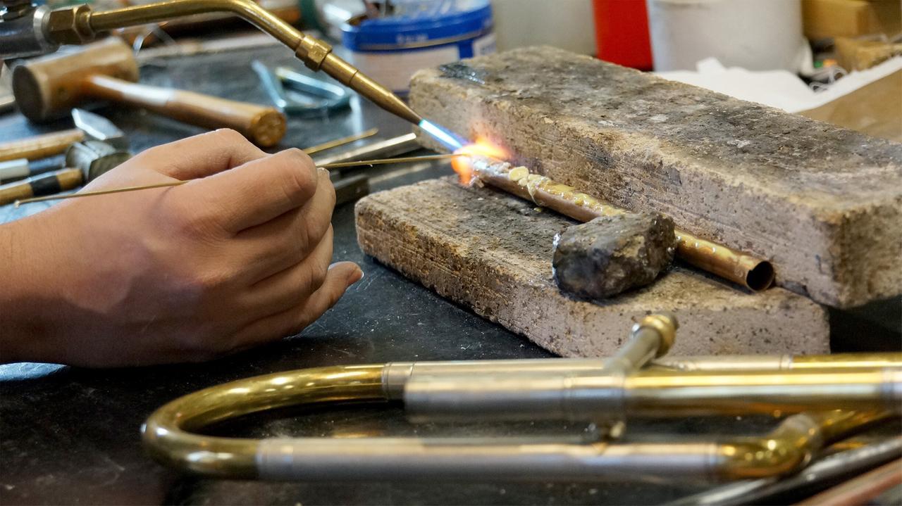 Das Bild zeigt einen Arbeitsplatz in einer Werkstatt für den Instrumentenbau. Im Vordergrund wird eine Trompete gelötet. Eine Hand hält ein Lötgerät, das eine blaue Flamme ausstößt und die Metallteile miteinander verbindet. Auf einem groben, grauen Arbeitsplatz aus Stein liegen verschiedene Werkzeuge wie Hammer und Zangen sowie andere metallene Teile. Eine kleine Metallstange ist auf einem Steinblock fixiert, und ein Stück Metall (wahrscheinlich Lötmaterial) ist ebenfalls sichtbar. Der Raum wirkt gut beleuchtet und der Fokus liegt klar auf dem Lötvorgang.