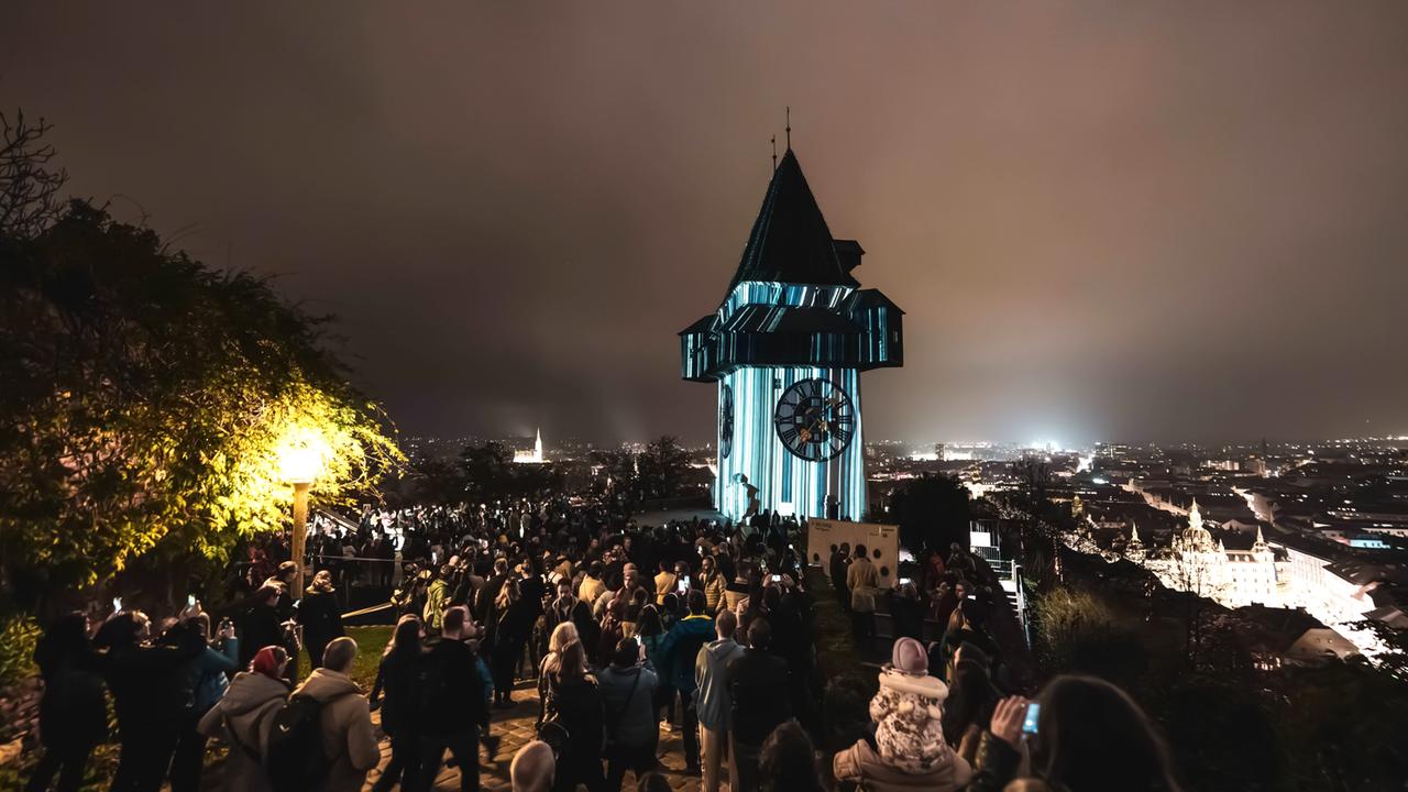 Das Bild zeigt eine große Menschenmenge, die sich um einen beleuchteten Uhrturm versammelt hat. Der Himmel ist dunkel und die Stadt im Hintergrund ist schwach erleuchtet. Smartphones werden gehalten.
