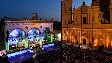 Bunt beleuchtet erstrahlt die Feldherrenhalle bei Klassik am Odeonsplatz, daneben die dezent angestrahlte Theatinerkirche, in der Mitte Publikum