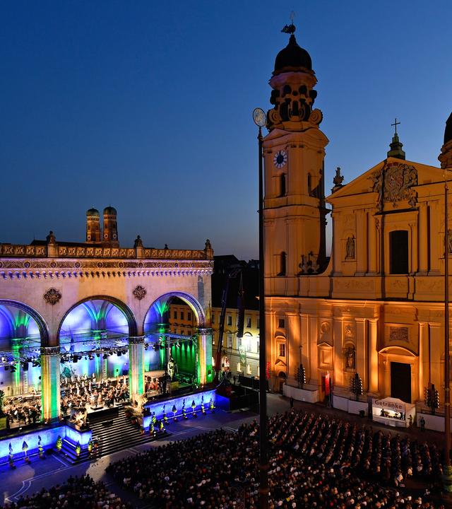 Bunt beleuchtet erstrahlt die Feldherrenhalle bei Klassik am Odeonsplatz, daneben die dezent angestrahlte Theatinerkirche, in der Mitte Publikum