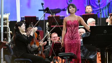 Yuja Wang trägt ein festliches pinkes Kleid. Sie steht links neben den Flügel und wird durch Applaus von den Musikern der Münchner Philharmonikern gefeiert. Die Orchstermusiker befinden sich im Hintergrund.