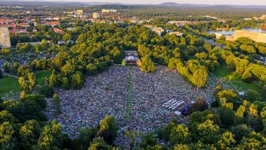 Eine große Fläche in einem Park bedeckt mit vielen bunten Objekten vor einer aufgebauten Bühne, aufgenommen aus der Vogelperspektive.